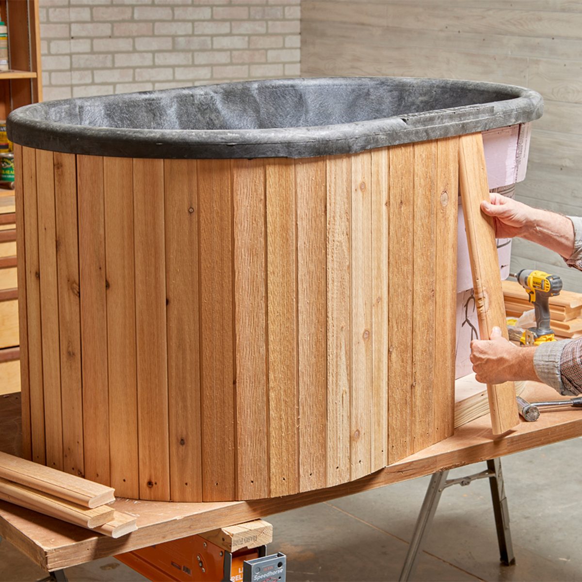 A wooden structure is being assembled by a person, who is fitting a plank into a round, partially constructed container in a workshop setting.