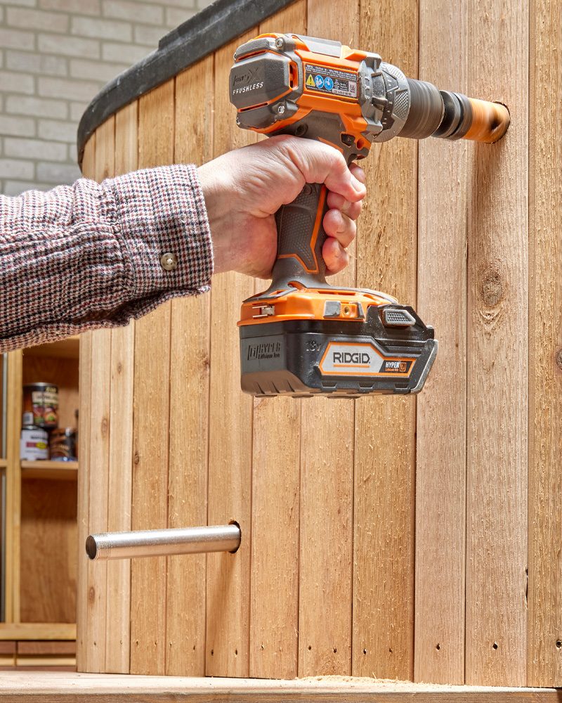 A person operates a drill, creating a hole in a wooden wall, while a metal pipe extends through the wall in a workshop setting.