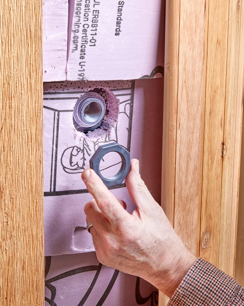 A hand holds a metal nut, positioning it over a plastic fitting embedded in insulated wall panels beside wooden framing.