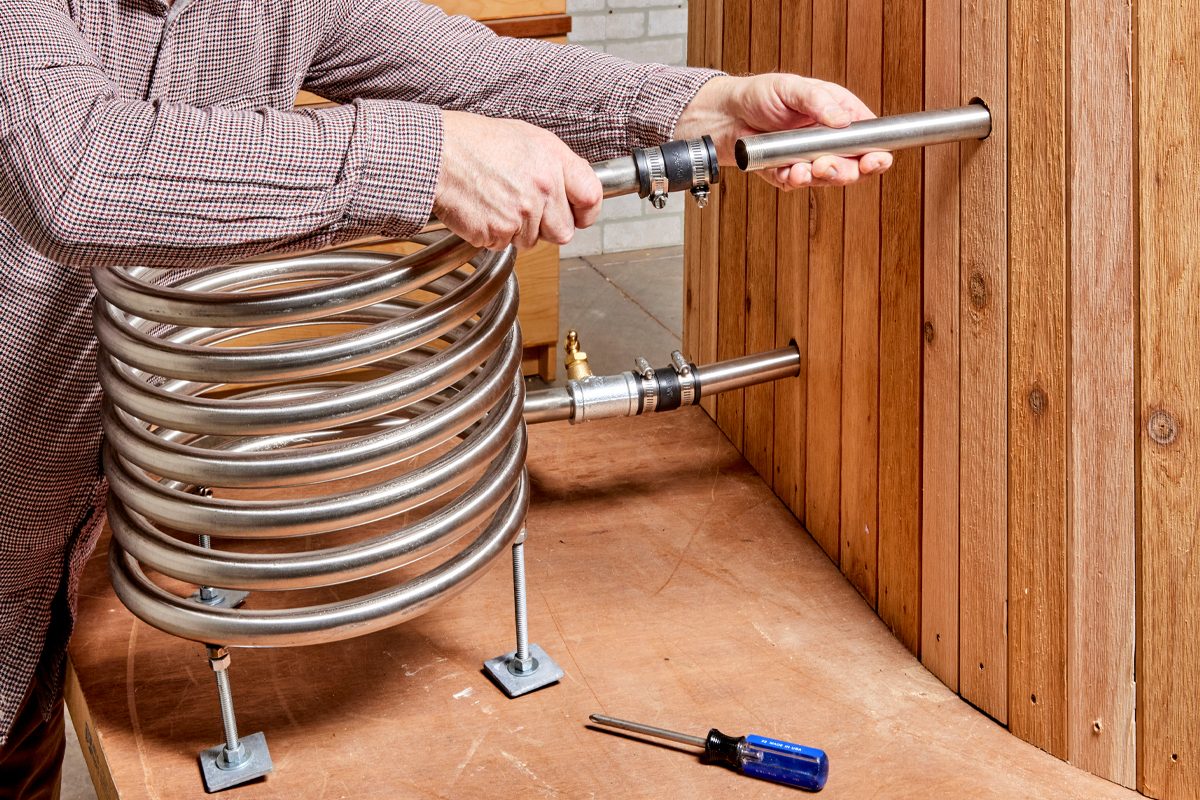 A person uses a pipe to connect a coiled metal structure to a wooden wall, surrounded by tools on a work surface.