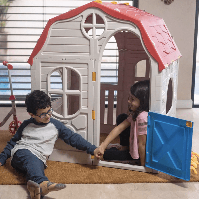 Two children sit on a floor by a plastic playhouse, smiling and holding hands through an open door, with playful toys nearby.