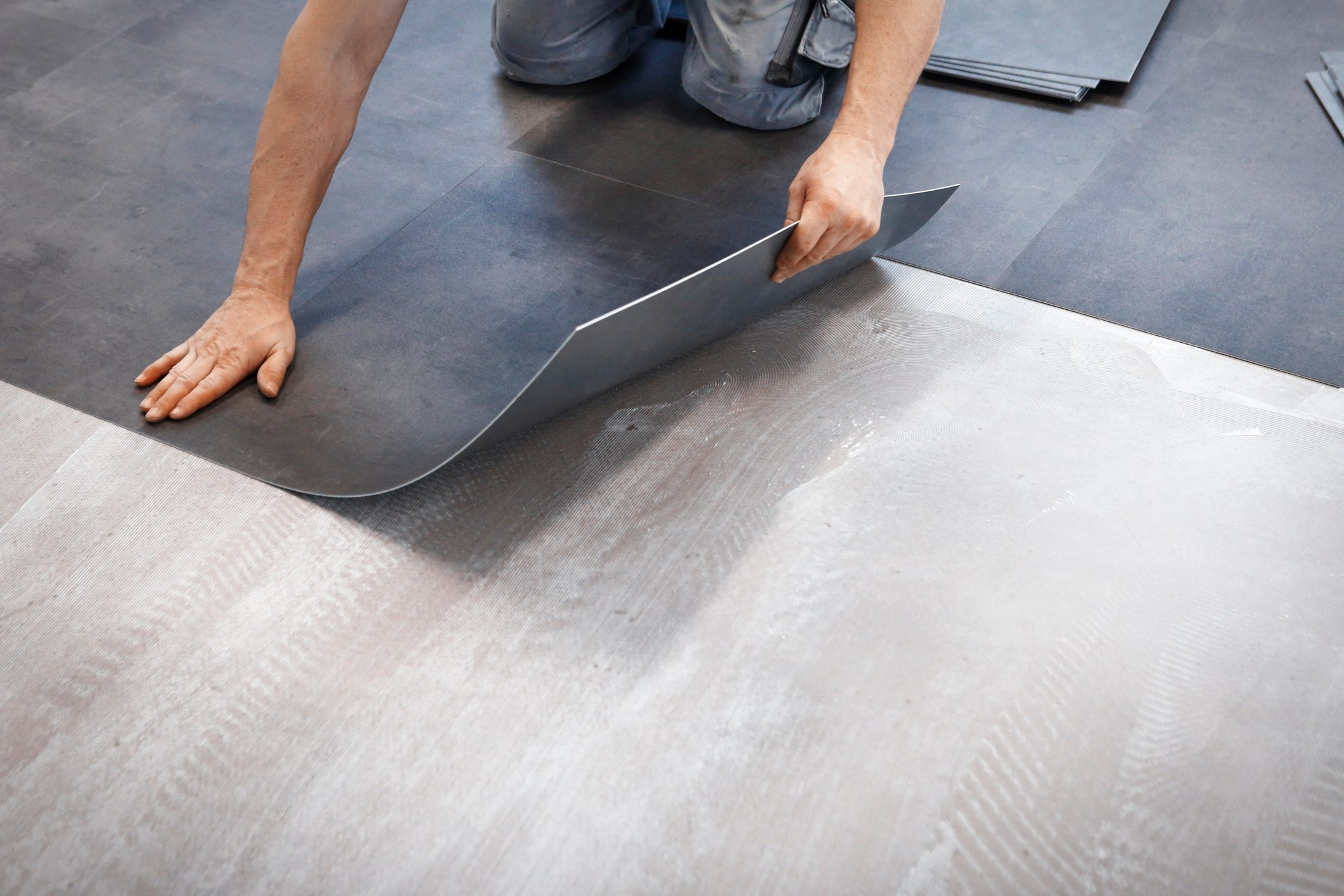 A person positions a dark flooring tile above a lighter surface, preparing to lay it down in a room undergoing renovation.