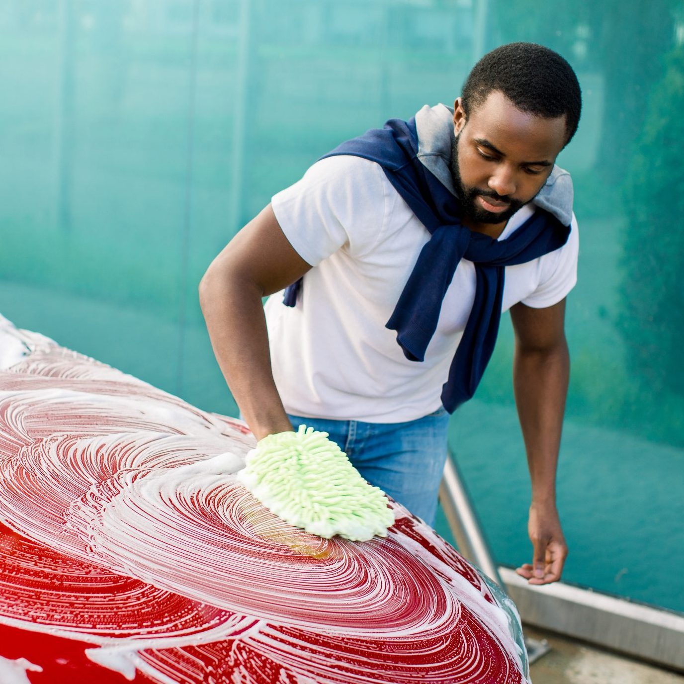 A man washes a car using a green microfiber cloth, applying soap in circular motions on its red surface, with a blue-green background visible.