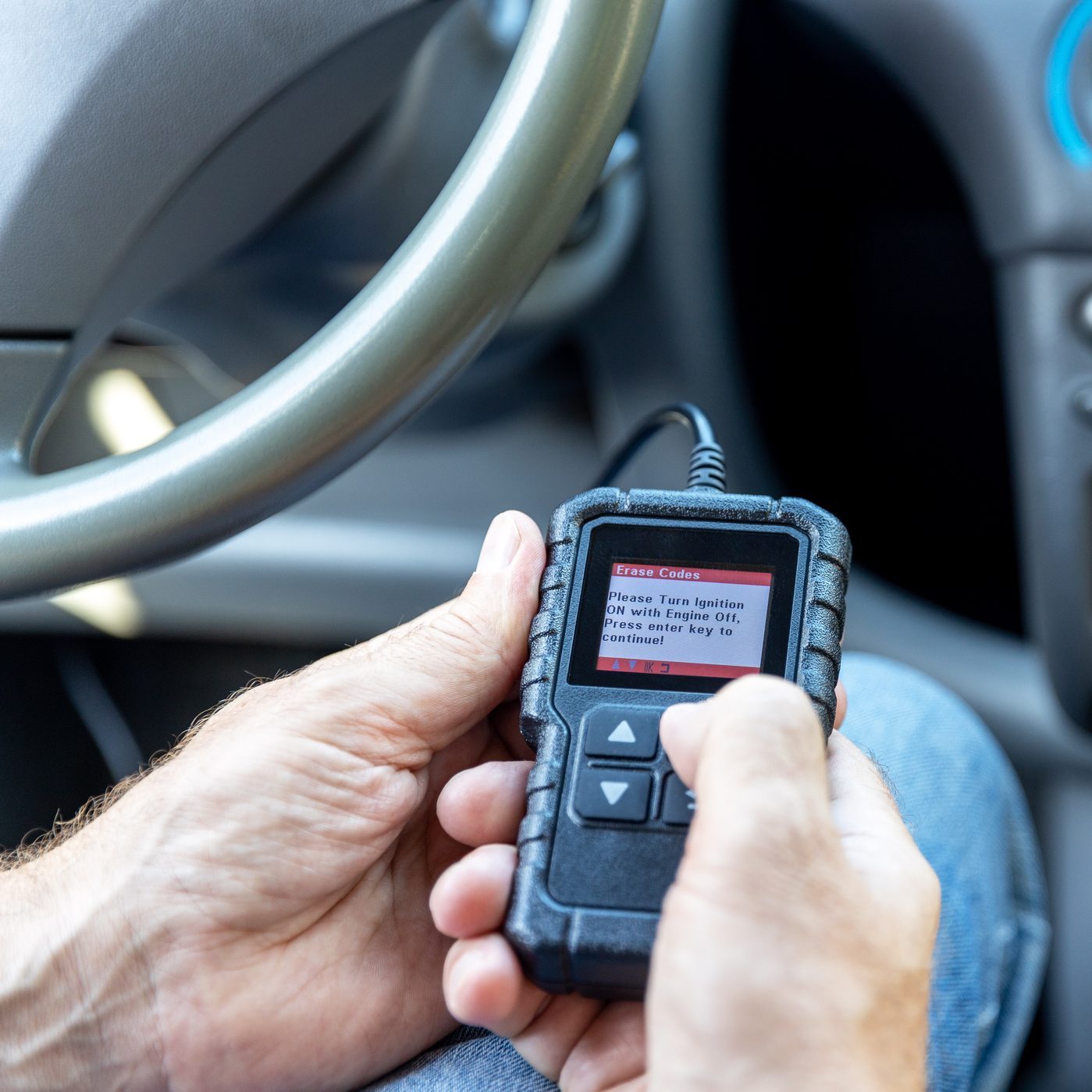 A hand holds a diagnostic tool displaying instructions to turn on the vehicle's ignition while sitting in a car with a steering wheel in view.