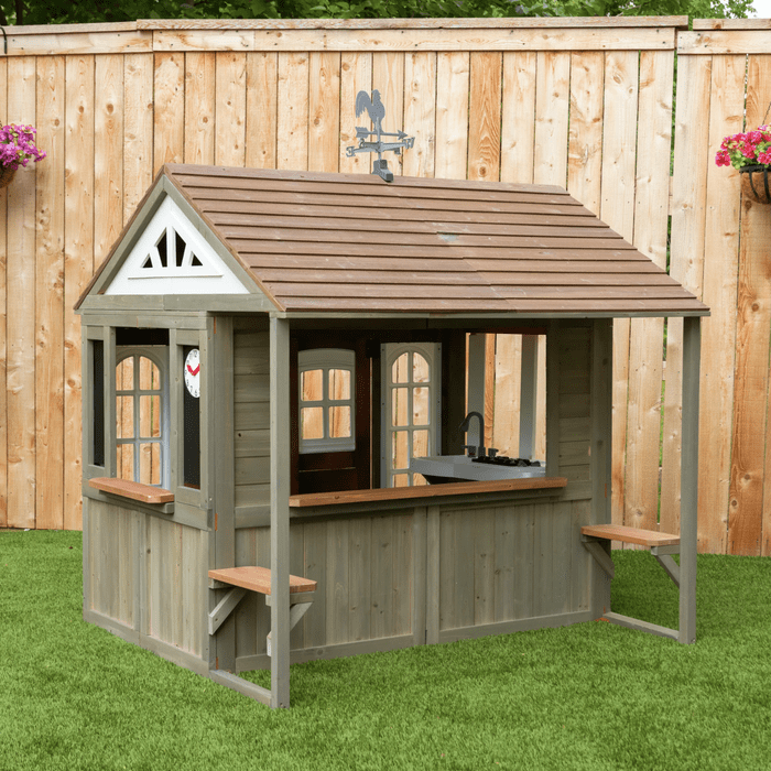 A wooden playhouse with a sloped roof and multiple windows. It features an outdoor countertop with a sink and is surrounded by green grass and a wooden fence.