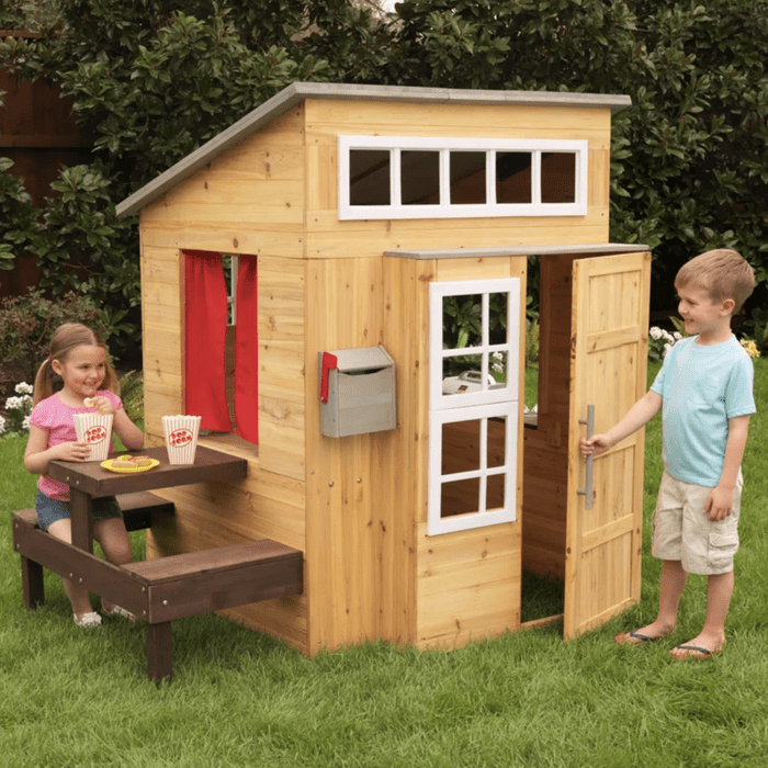 A wooden playhouse stands in a garden. A girl eats snacks at a table while a boy opens the door, inviting play. Green grass surrounds them.