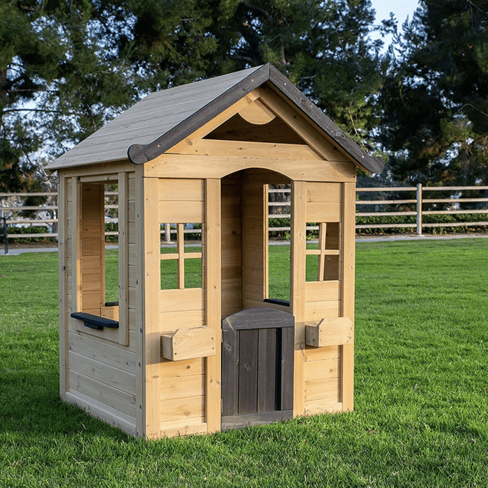 A wooden playhouse with a peaked roof stands on green grass, featuring open windows, a door, and a natural outdoor setting in the background.