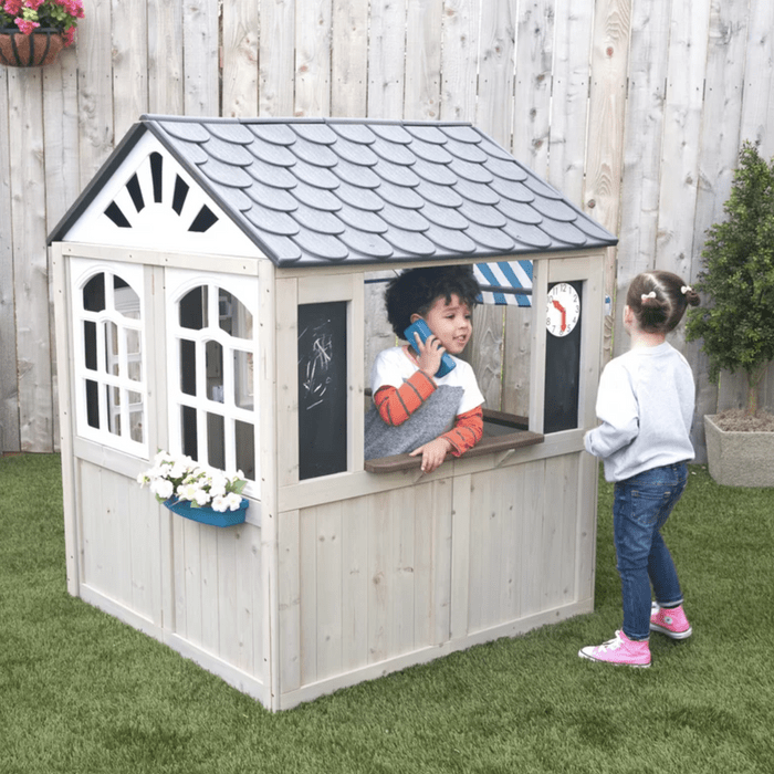Two children interact playfully at a wooden playhouse; one talks on a phone from a window while the other stands nearby, smiling.