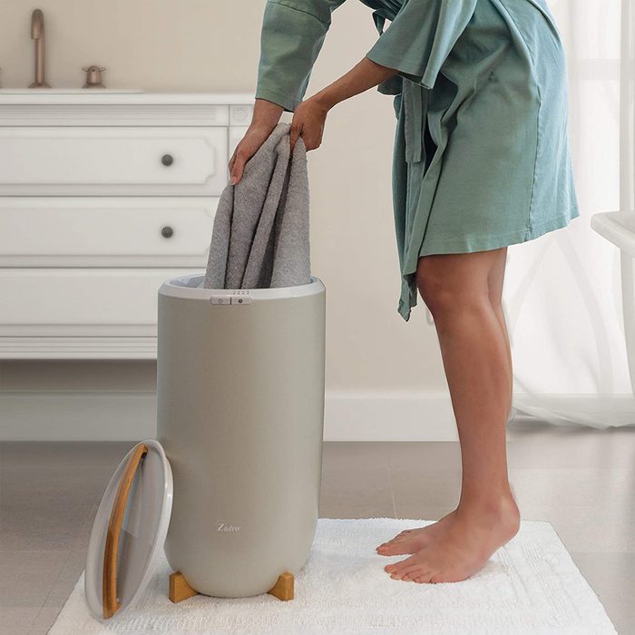 A person in a robe places a gray towel into a modern laundry hamper in a bright bathroom, standing on a white mat.