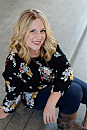 A woman with wavy hair smiles while sitting on a wooden floor, wearing a floral shirt and jeans, with a light background creating a warm atmosphere.