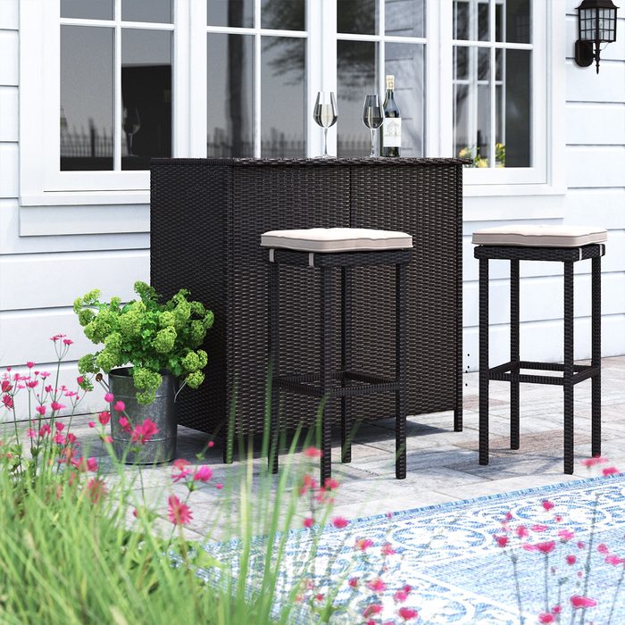 A dark wicker bar with two cushioned stools sits on a patio surrounded by pink flowers and a green plant, near a windowed wall.