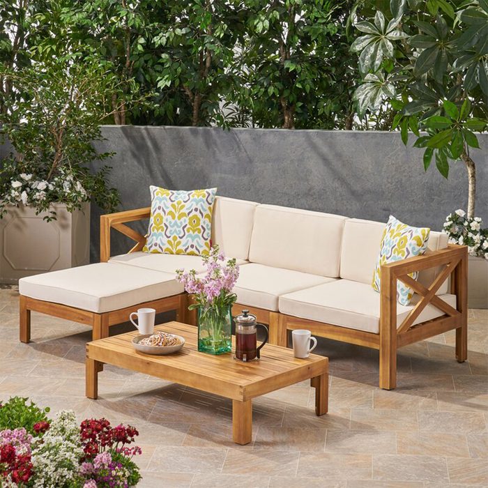 A wooden outdoor seating arrangement with cream cushions and decorative pillows surrounds a coffee table, featuring snacks and flowers in a lush, green setting.