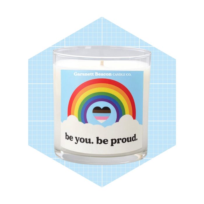 A candle displays a colorful rainbow and heart design, promoting a message of self-acceptance against a light blue grid background.