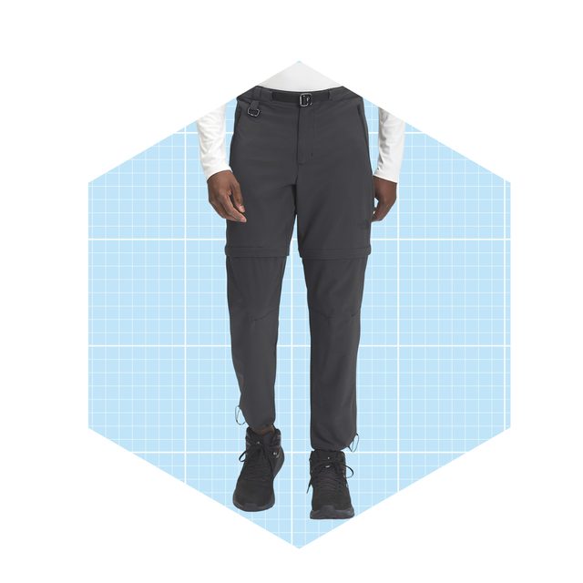 Dark cargo pants with zippers are worn by a person standing against a light blue grid background. The person is ready for outdoor activities.