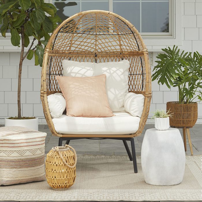 A woven chair with a cushion sits on a patterned rug, surrounded by decorative plants and tables in a bright, inviting outdoor space.