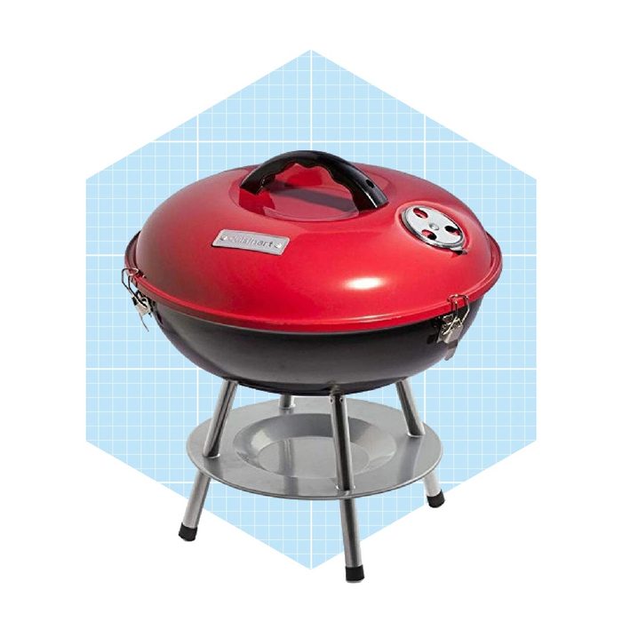 A round, red and black portable grill sits on a three-legged stand, designed for outdoor cooking, with a vented lid to manage heat.