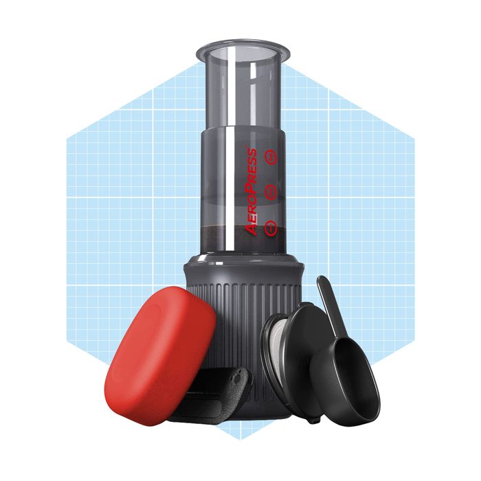 An AeroPress coffee maker sits upright, with coffee grounds inside. Accessories, including a red lid and black scoop, surround it on a blue grid background.