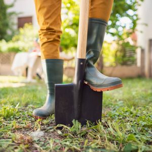 A person in green boots stands on a shovel in a garden, preparing to dig into the grass surrounded by greenery and a house in the background.