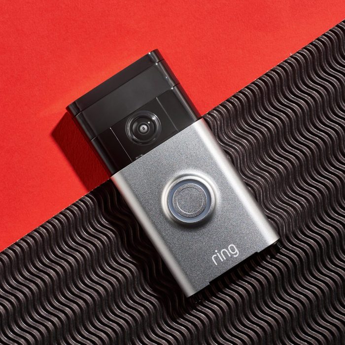 A video doorbell rests on a textured surface, capturing its sleek design. The background features a vibrant red color, contrasting with the doorbell's metallic finish.