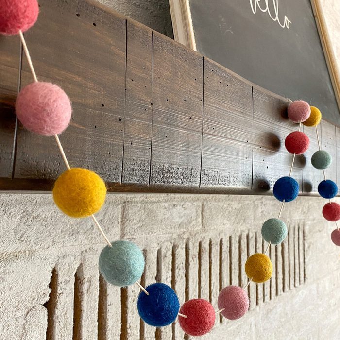 A colorful felt ball garland hangs from a wooden shelf against a textured wall, adding a playful touch to the interior decor.