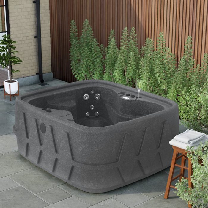 A dark hot tub sits on stone tiles, surrounded by greenery and a wooden wall, with a glass and towel on a nearby stool.