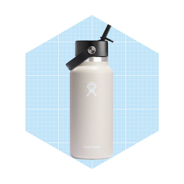 A stainless steel water bottle stands upright, featuring a black lid and a carrying handle, set against a light blue, grid-patterned background.
