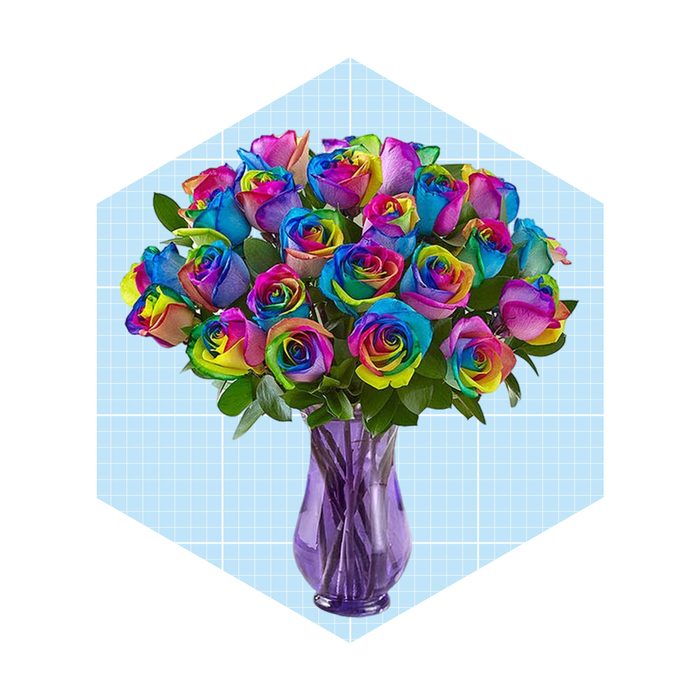 A vibrant bouquet of multicolored roses fills a purple vase, set against a light blue grid background. Lush green leaves complement the striking blooms.