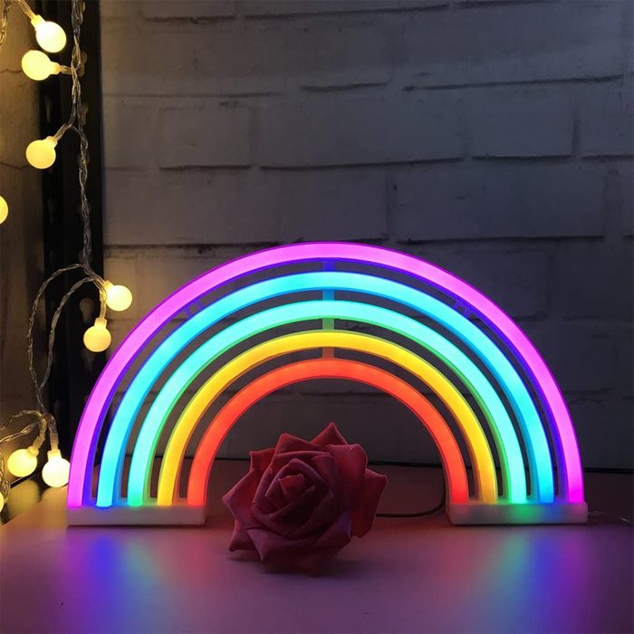 A colorful neon rainbow lamp illuminates a surface, complemented by a pink rose nearby, with soft glowing lights in the background.