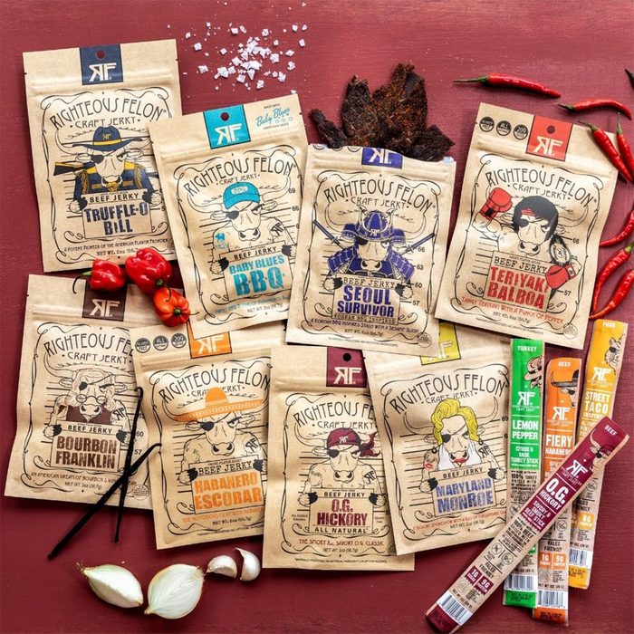 Various beef jerky packages, depicting unique characters, are arranged attractively with some red chili peppers and garlic cloves on a wooden surface.