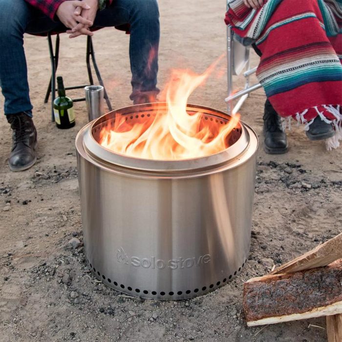 A metallic fire pit burns brightly on sandy ground, surrounded by seated individuals, with a nearby bottle and split wood visible.