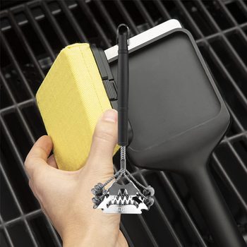 A hand holds a cleaning tool with a metal scraper and a yellow sponge, positioned near a grill's cooking surface.