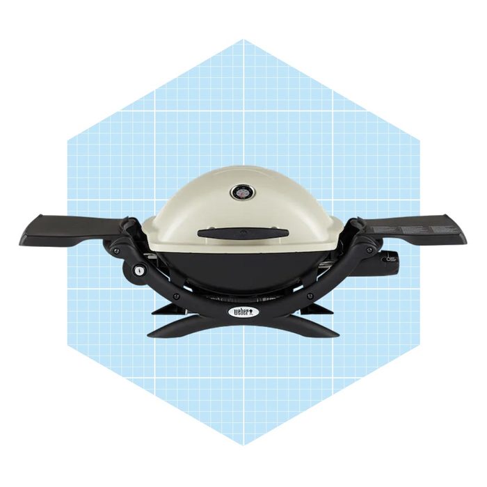 A compact gas grill with a rounded lid and foldable side tables is displayed against a light blue, grid-patterned background.