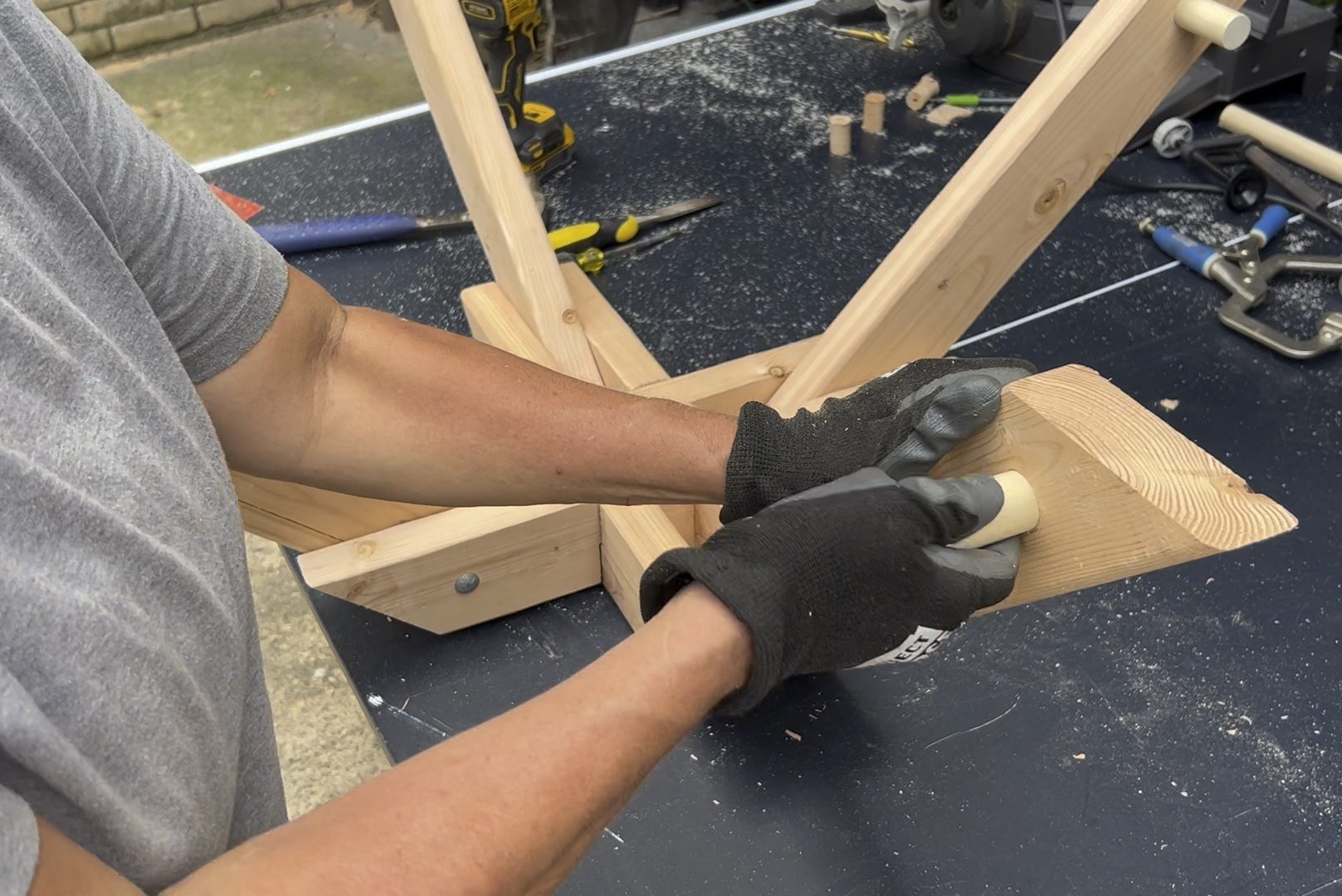 A person wearing gloves secures a wooden piece to a structure on a workbench, surrounded by tools and wood shavings outdoors.