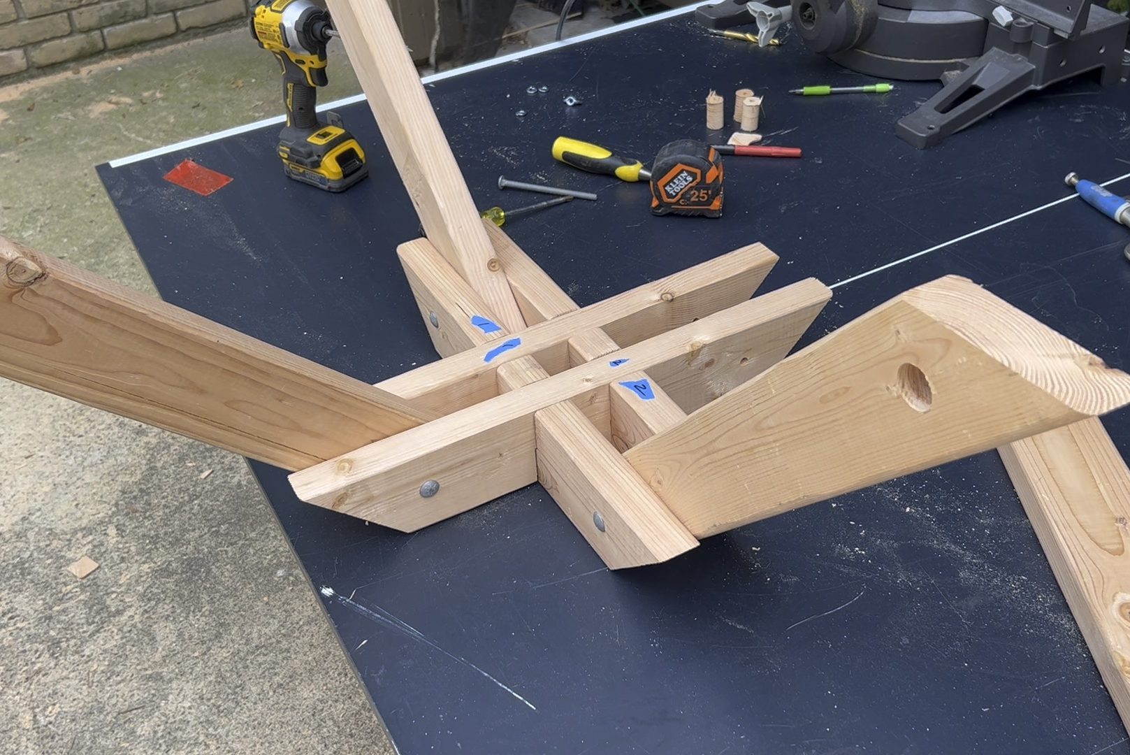 Wooden components are assembled on a black work surface, forming a structure with angled pieces. Tools and small parts are scattered nearby.