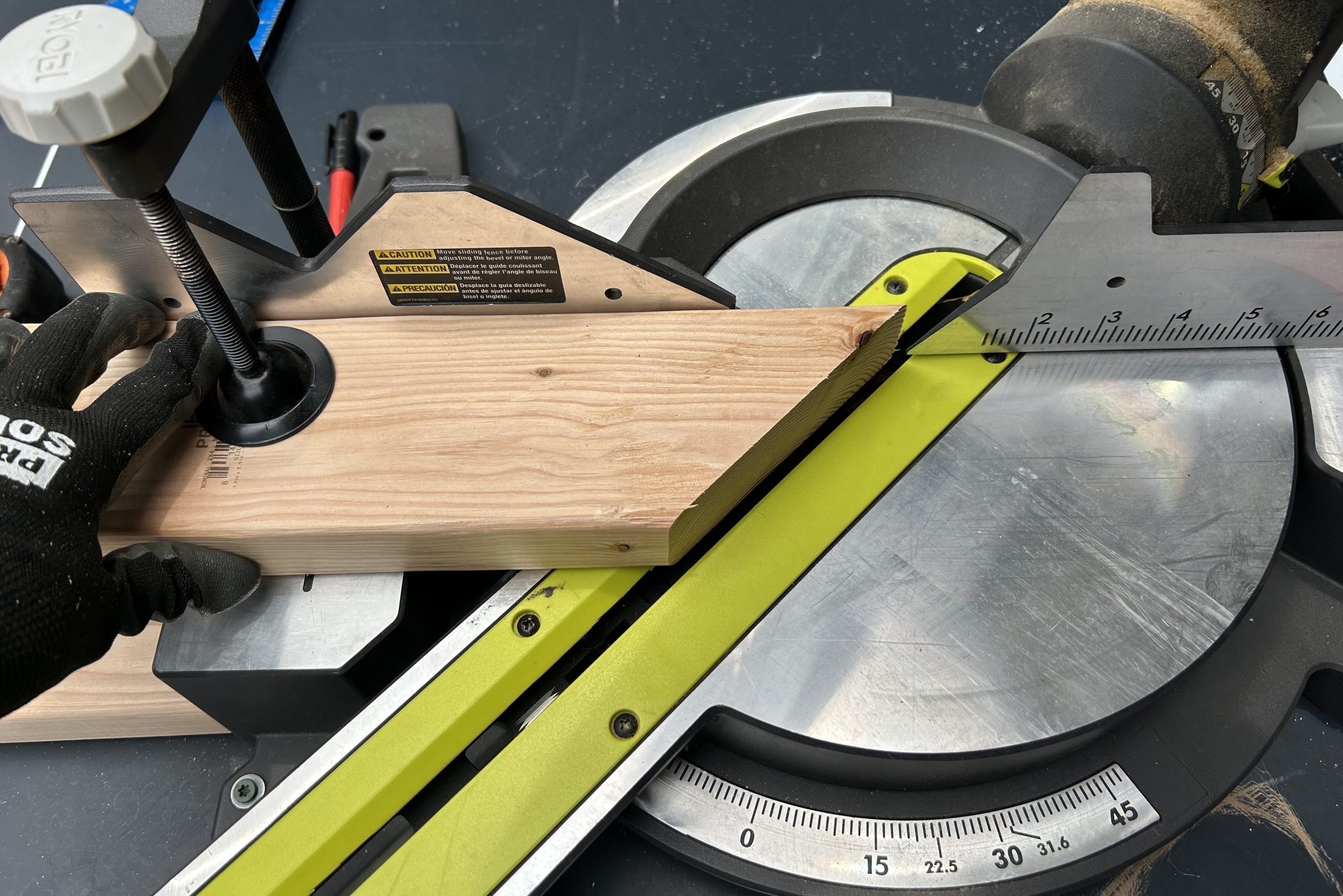 A wooden board is secured to a miter saw, preparing for a cut. The surrounding environment features various tools and equipment on a work surface.