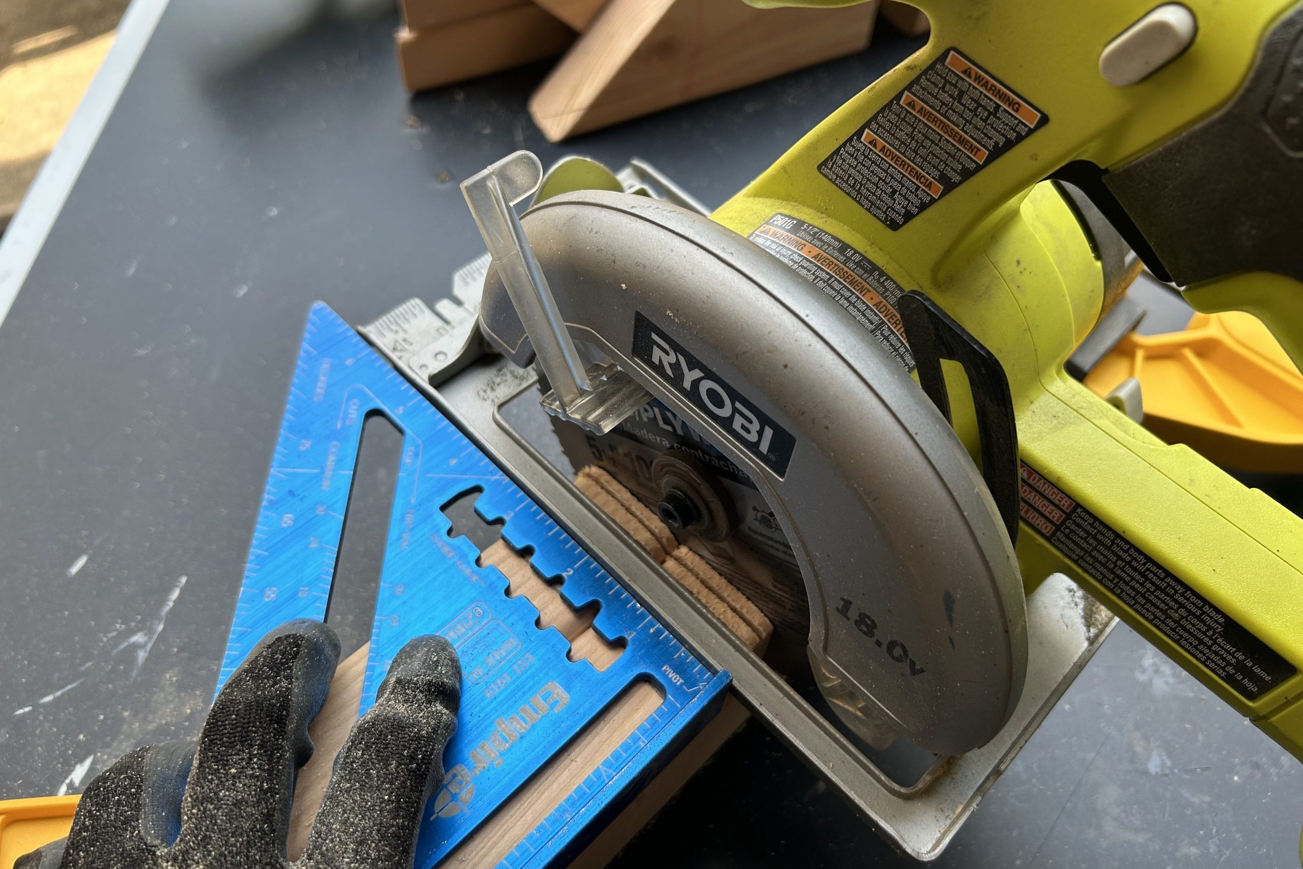 A circular saw is positioned on a work surface, cutting through wood while held steady by a blue triangular square and gloved hand, surrounded by dust.