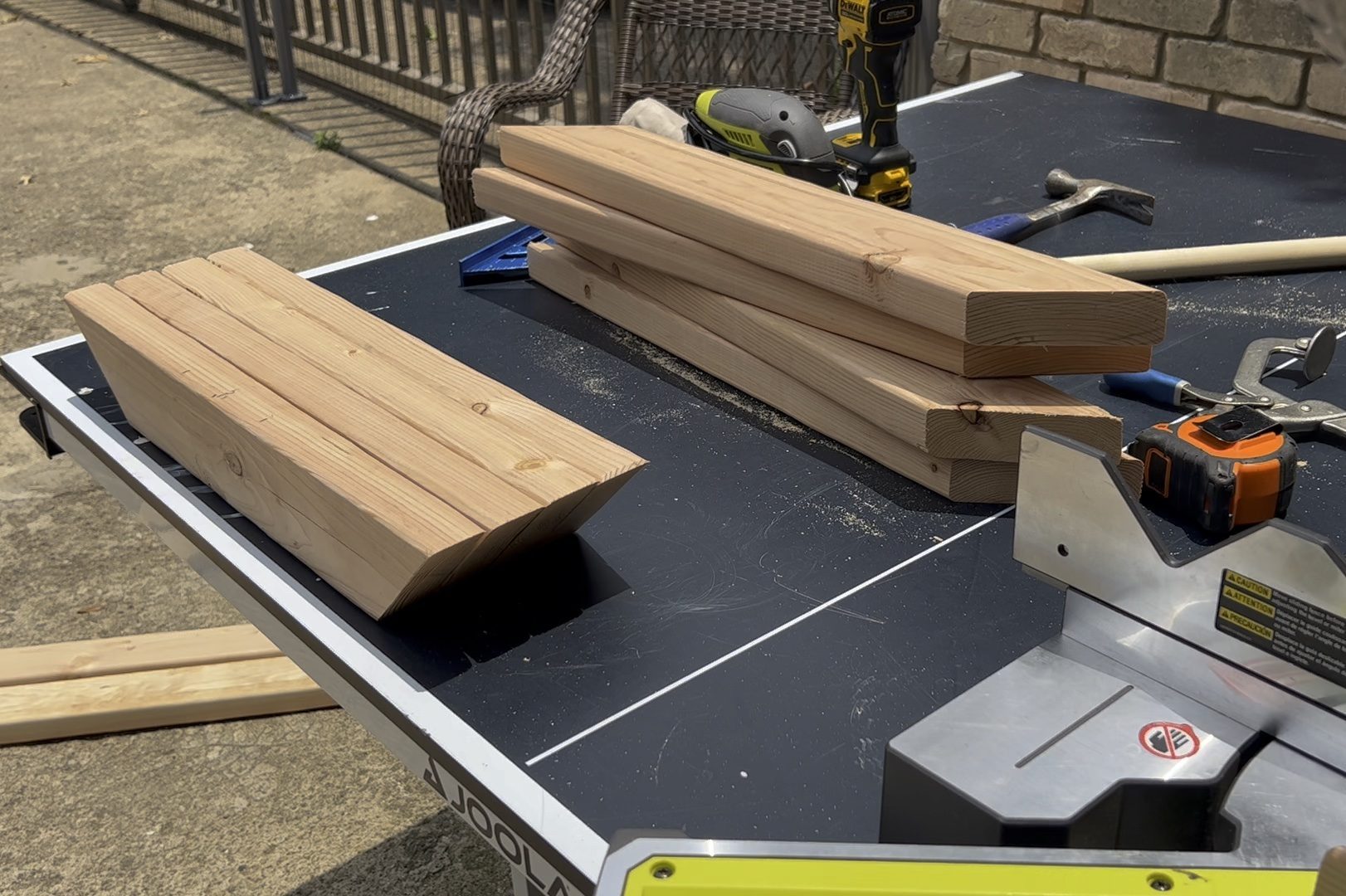 Wooden planks rest on a black table, surrounded by tools. Sunlight illuminates the scene, suggesting an outdoor woodworking project in progress.