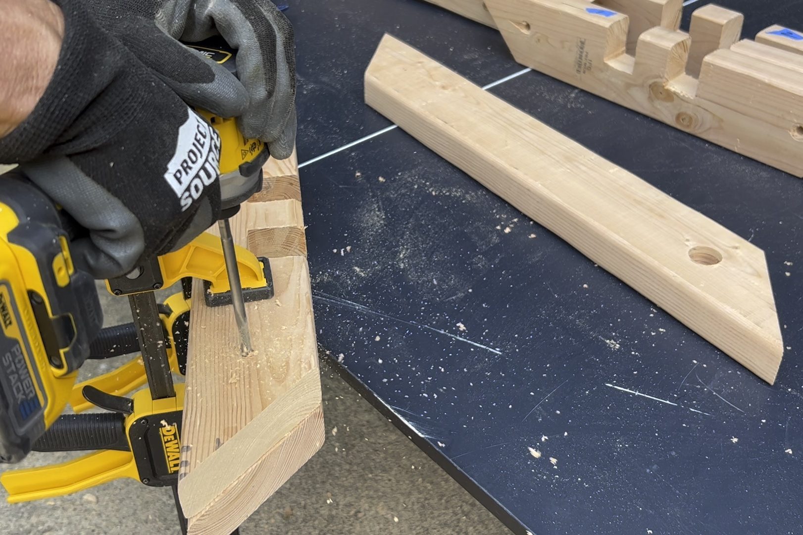 A power drill is drilling into a wooden plank, resting on a black table, surrounded by wood shavings and additional wooden pieces.