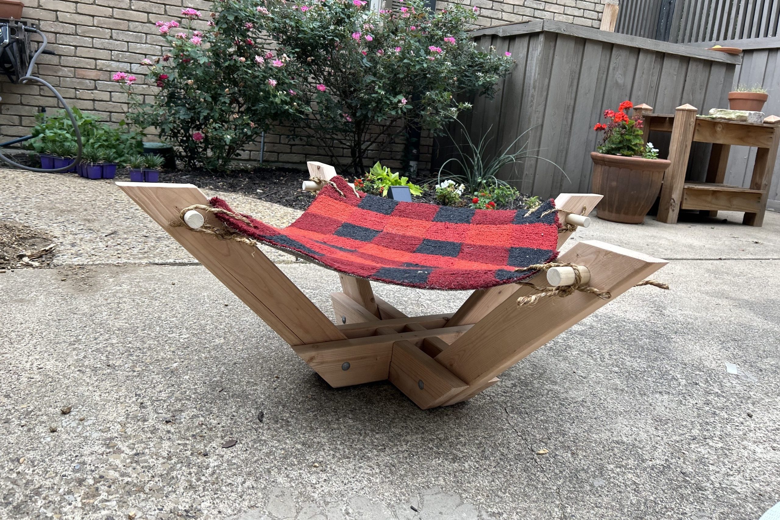A handmade wooden chair with a red and black checkered fabric seat rests on a concrete surface, surrounded by flowering plants and garden decor.