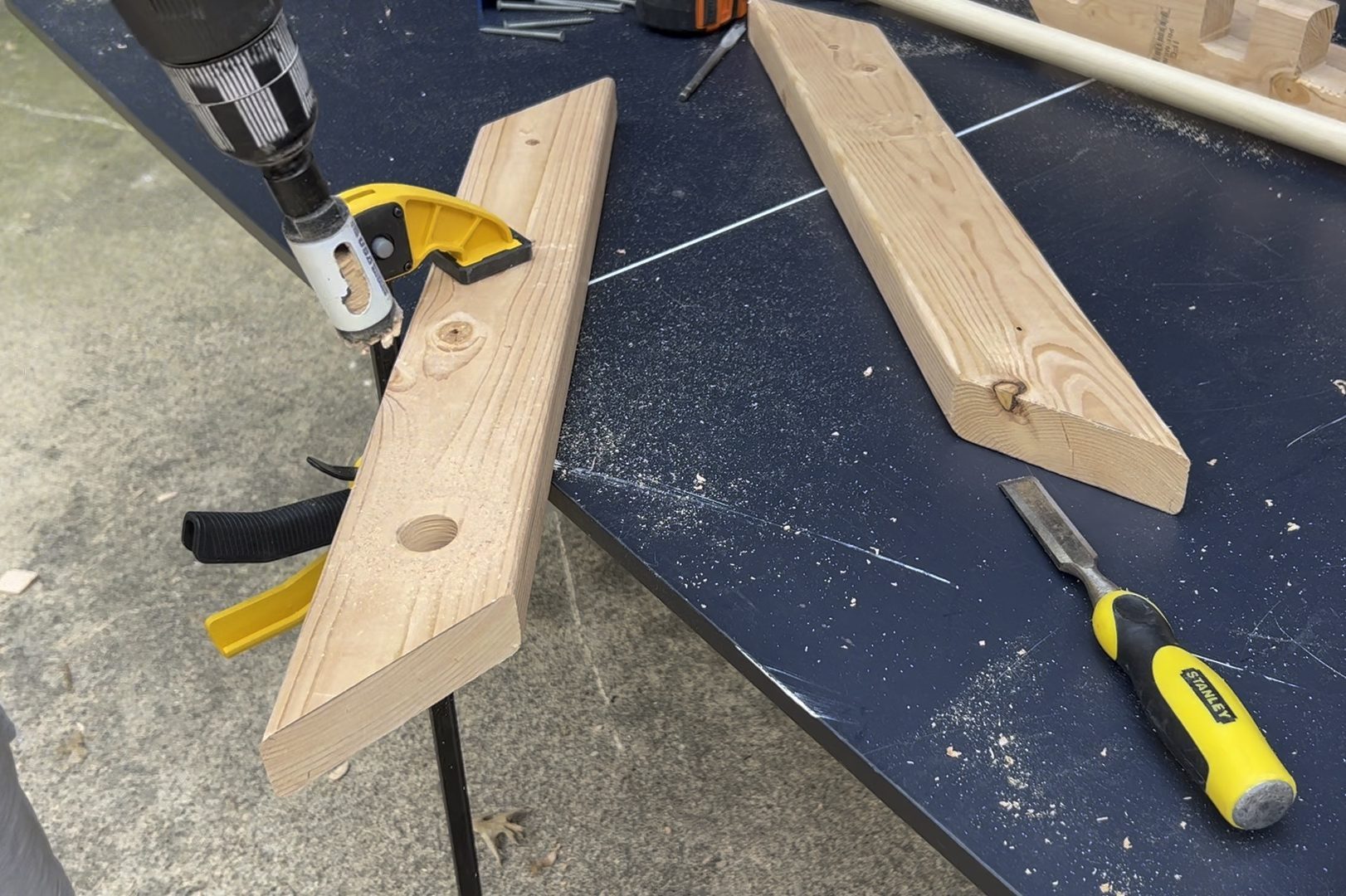 A drill is cutting through a wooden plank on a worktable, surrounded by sawdust and tools, including another plank and a chisel.