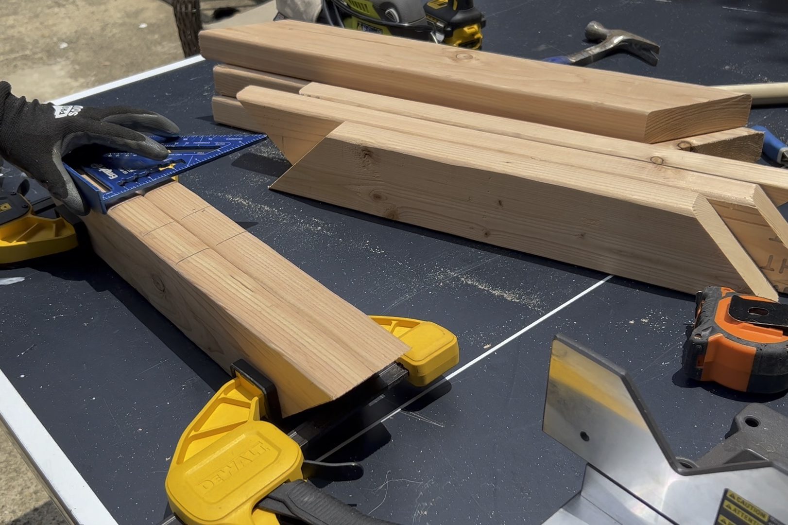 A hand with a gloved finger adjusts a square tool against a wooden beam, while multiple raw wooden pieces rest on a black work surface outdoors.