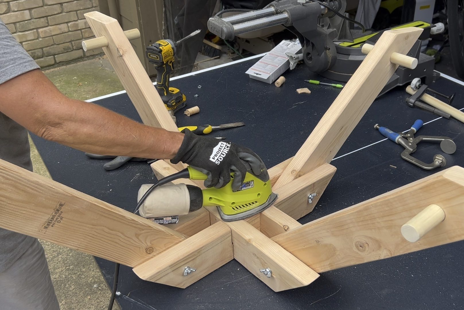 A hand operates a sanding tool on wooden beams arranged in a star formation, with various tools and materials scattered across a dark work surface.