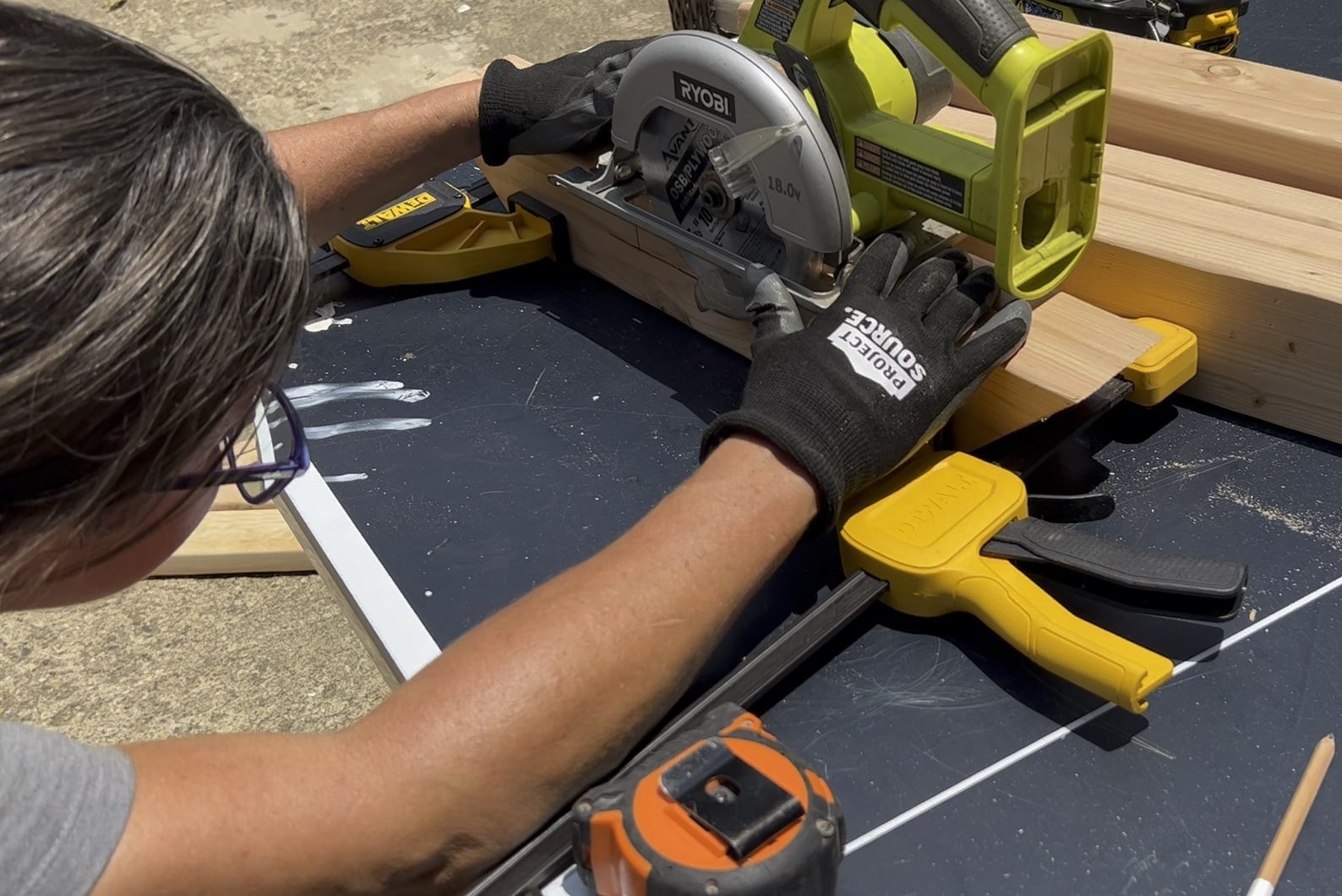 A person uses a circular saw to cut wood, positioned on a black work surface outdoors, secured with clamps and surrounded by tools and materials.