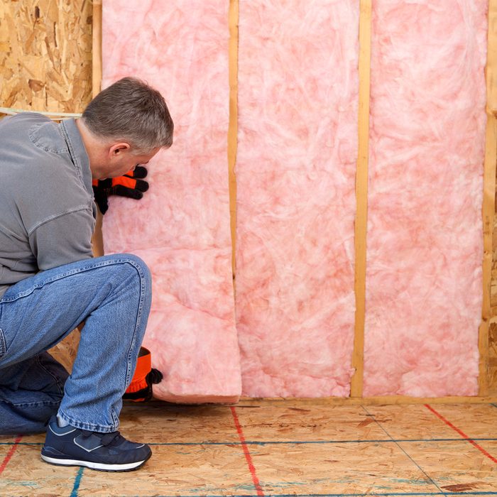 A person installs pink insulation between wooden studs in a room. They're crouched down, focusing on the material in a partially completed space.