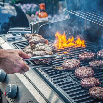 A hand uses tongs to flip meat on a grill, where flames rise and smoke billows. The setting suggests a casual outdoor cooking environment.