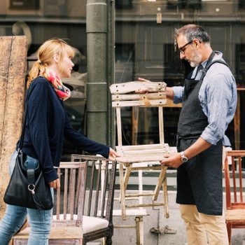 A woman examines a wooden chair held by a man wearing an apron, surrounded by various other chairs outside a shop.
