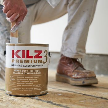A painter kneels, stirring a can of primer with a brush on a wooden floor, surrounded by an unfinished room. Brown work boots are visible.