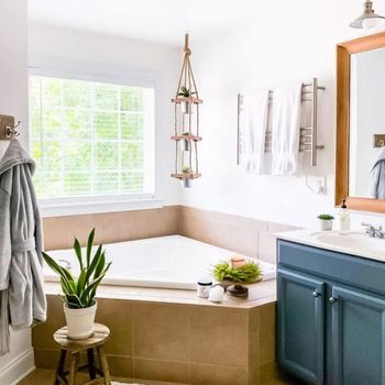 A corner bathtub sits in a bright bathroom, surrounded by plants, a stool, and hanging towels, while natural light filters through a window.