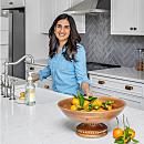 A woman in a blue shirt stands by a kitchen sink, smiling while preparing food beside a bowl of fresh fruits on a white countertop.