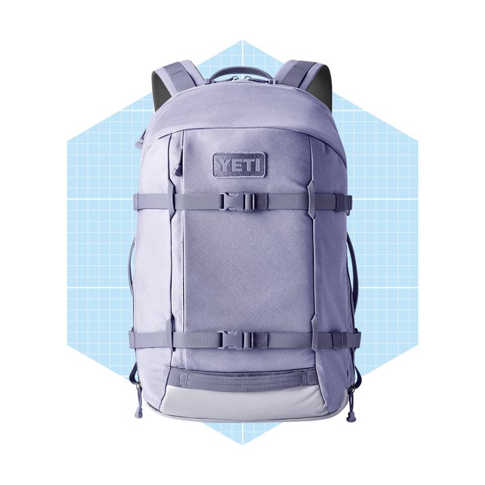 A light purple backpack with straps and buckles stands upright against a light blue geometric background, emphasizing its design and features.